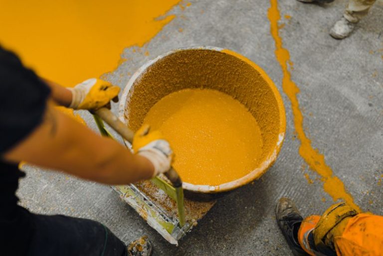Worker pouring mixed mustard-yellow epoxy resin from a bucket onto concrete